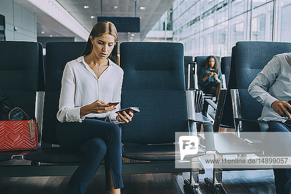 Junge Geschäftsfrau sendet Textnachrichten über ein Smartphone  während sie mit ihrem Pass im Wartezimmer des Flughafens sitzt