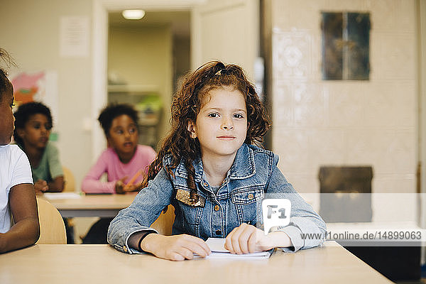 Porträt eines lächelnden Schulmädchens  das mit Freunden am Schreibtisch im Klassenzimmer sitzt