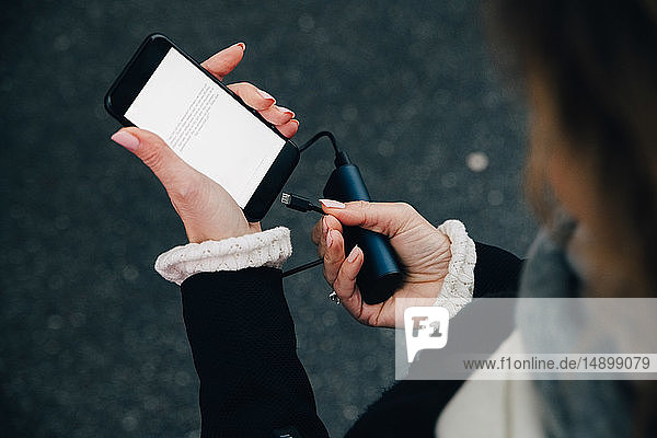 Mittelsektion einer Geschäftsfrau  die ein Ladegerät in ein Mobiltelefon einsteckt  während sie auf der Straße steht