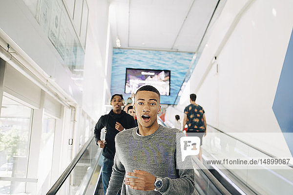 Portrait of shocked teenage boy moving down on escalator with friend in background at shopping mall
