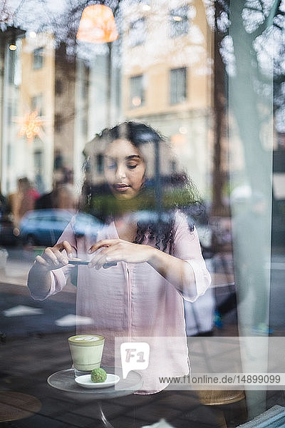 Junge einflussreiche Frau fotografiert Tee und Snack auf dem Tisch durch ein Smartphone  das durch ein Fenster gesehen wird