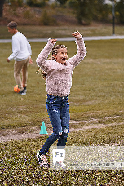 Lächelnde Schwester mit erhobenen Armen auf Grasfeld stehend  während der Bruder im Hintergrund im Park Fussball spielt