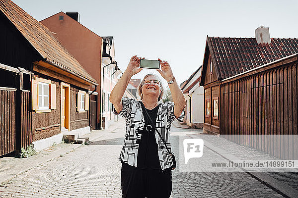 Lächelnde ältere Frau fotografiert  während sie auf der Straße in der Stadt vor klarem Himmel steht
