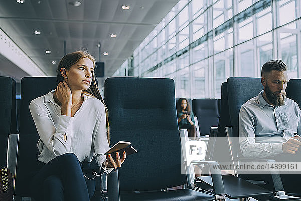 Nachdenkliche junge Geschäftsfrau schaut weg  während sie neben einem Kollegen im Wartebereich des Flughafens sitzt