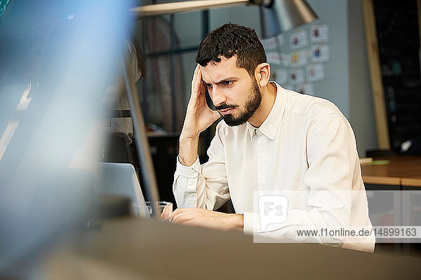 Angespannter Geschäftsmann schaut auf Laptop  während er im Kreativbüro sitzt