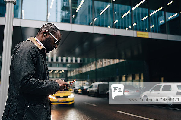 Side view of businessman using smart phone while standing on road against building in city