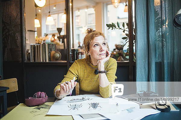 Nachdenkliche reife Illustratorin schaut weg  während sie mit Zeichenpapier am Schreibtisch im Kreativbüro sitzt