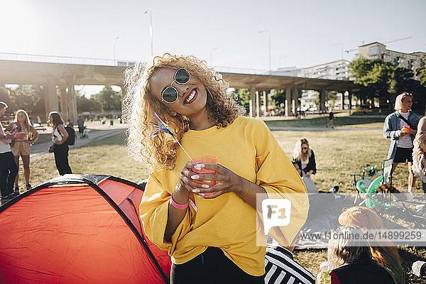Porträt einer lächelnden Frau mit Getränk  die bei einer Musikveranstaltung im Sommer gegen Freunde steht
