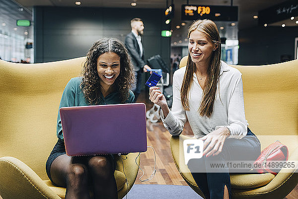 Lächelnde multiethnische Kolleginnen diskutieren über Laptop und Kreditkarte  während sie in der Flughafen-Lobby sitzen