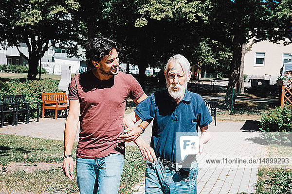 Smiling male caretaker looking while walking arm in arm with senior man at nursing home
