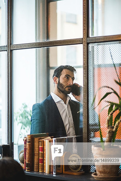 Selbstbewusster Geschäftsmann spricht am Smartphone  gesehen durch Glaswand im Kreativbüro