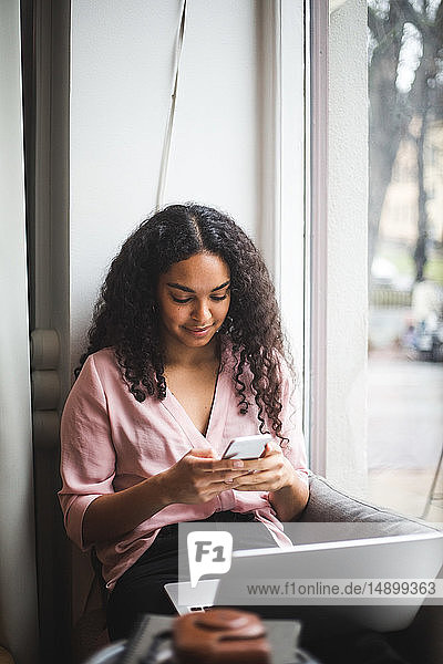 Junge berufstätige Frau  die im Büro auf dem Fensterbrett sitzt und Textnachrichten per Smartphone verschickt