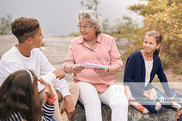 Glückliche Großmutter schenkt Enkel  während sie mit Enkelinnen im Park beim Picknick sitzt