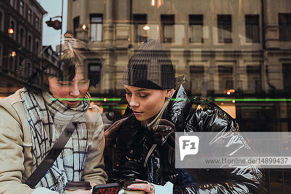 Junge Frau trägt warme Kleidung  während sie eine Freundin im Café durch ein Glasfenster betrachtet  die ein Mobiltelefon benutzt