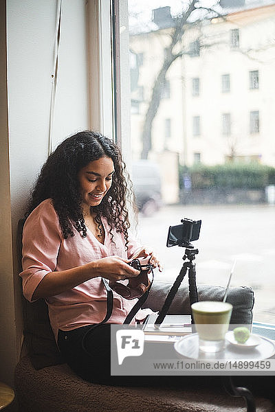 Lächelnde junge einflussreiche Frau hält Kamera am Fenster im Kreativbüro