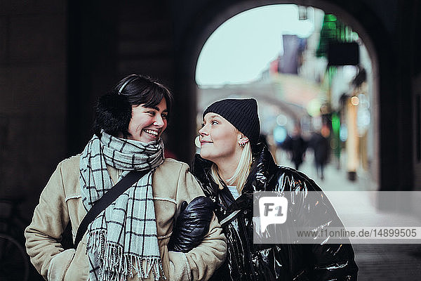 Smiling young female friends looking each other face to face while standing on street in city during winter