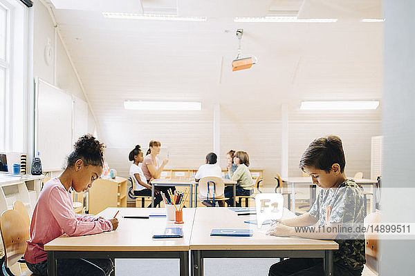 Boy and girl writing at desk while friends learning with student in background