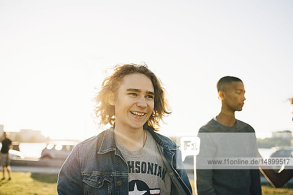 Lächelnder junger Mann  der wegschaut  während sein Freund im Hintergrund vor dem klaren Himmel an einem sonnigen Tag steht