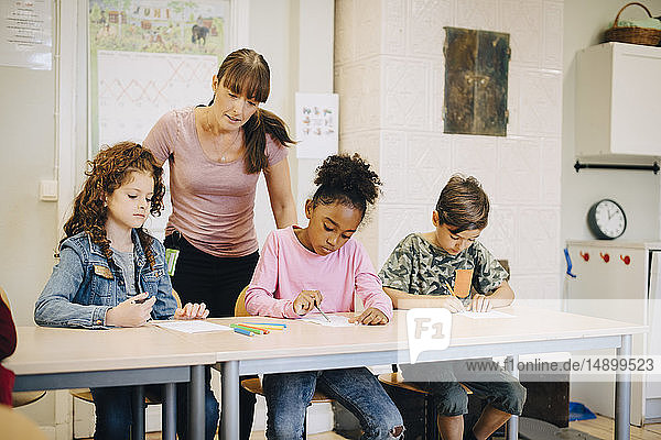 Lehrer betrachtet Schüler  die in der Grundschule am Schreibtisch schreiben