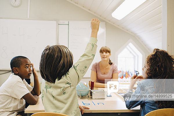 Schoolboy raising hand while answering teacher in classroom at school