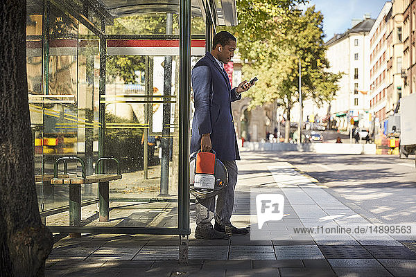 Seitenansicht in voller Länge eines jungen männlichen Pendlers  der ein Smartphone benutzt  während er ein elektrisches Einrad an einer Bushaltestelle in der Stadt hält