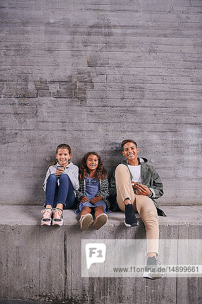 Full length portrait of happy siblings sitting side by side against wall at playground