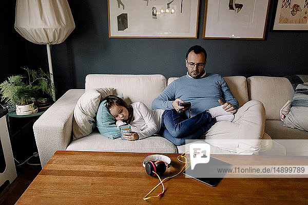 Father and daughter using mobile phones on couch in living room at home