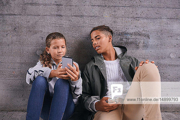 Sister showing her mobile phone to brother while sitting against wall