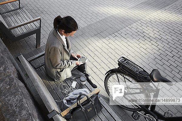 Schrägaufnahme einer Frau  die mit einem Smartphone im Internet surft  während sie in der Stadt auf einer Bank sitzt