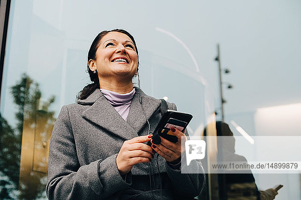 Niedriger Blickwinkel auf eine lächelnde Geschäftsfrau  die ein Mobiltelefon benutzt  während sie gegen ein Gebäude in der Stadt steht