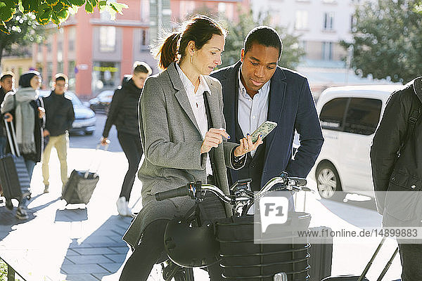 Eine reife Frau zeigt einem jungen Mann ein Smartphone  während sie in der Stadt mit einem Elektrofahrrad steht