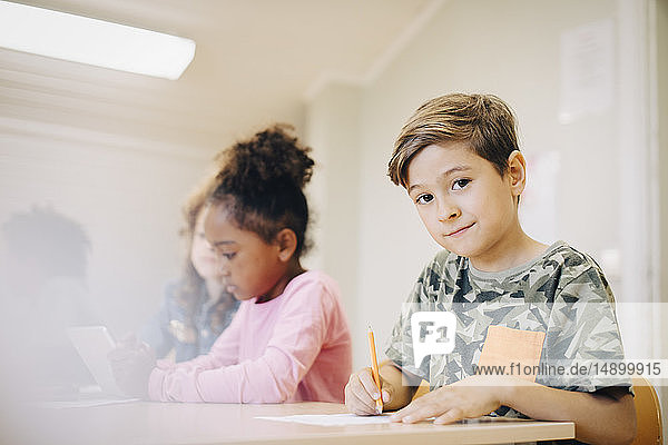 Portrait of boy sitting with friend writing on paper at desk in classroom