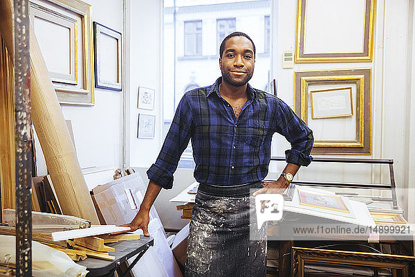 Porträt eines lächelnden jungen männlichen Künstlers  der mit der Hand auf der Hüfte in einem Rahmenwerkstatt steht