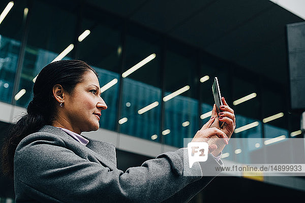 Seitenansicht einer Geschäftsfrau  die mit einem Smartphone fotografiert  während sie vor einem modernen Gebäude in der Stadt steht