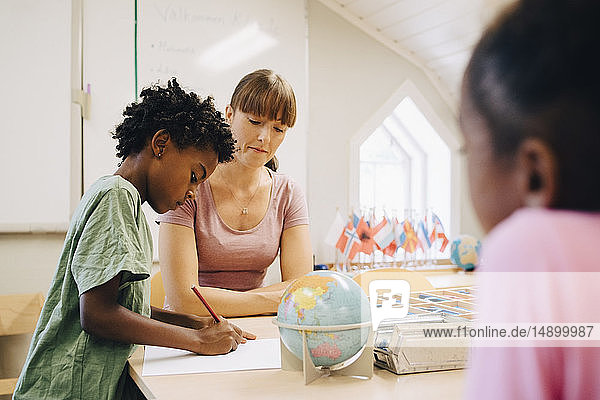 Teacher looking at boy writing on paper at table in classroom