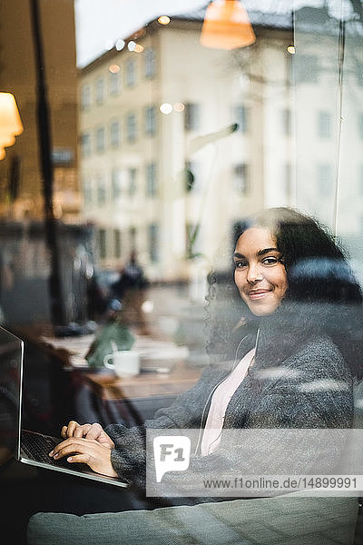Porträt einer lächelnden jungen Berufstätigen  die mit einem Laptop durch ein Fenster im Büro sitzt