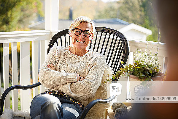 Bildnis einer fröhlichen reifen Frau mit verschränkten Armen auf der Veranda sitzend