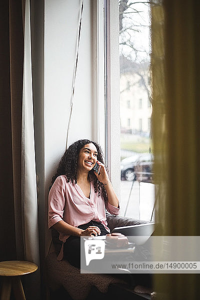 Lächelnde junge Berufstätige  die im Büro auf dem Fensterbrett sitzt und durch ein Smartphone spricht