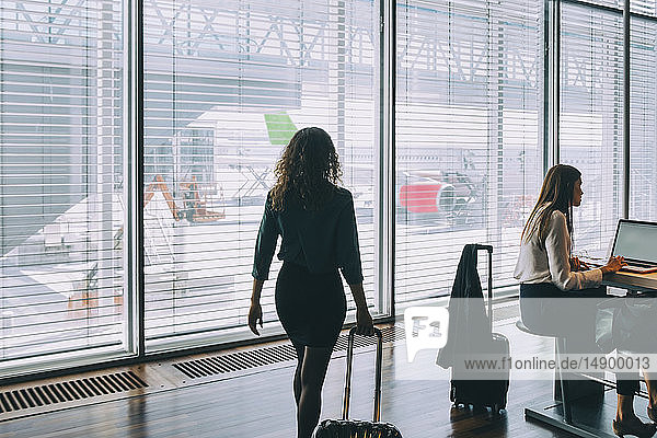 Rückansicht einer mit Gepäck gehenden Geschäftsfrau von einer im Abflugbereich des Flughafens sitzenden Kollegin