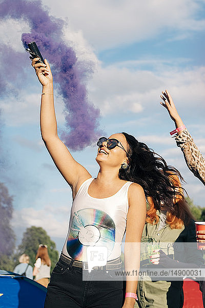 Fröhliche Frau hält Notfall-Fackel mit lila Rauch in der Hand  während sie mit Freunden ein Musikkonzert genießt