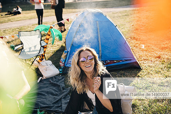 Hochwinkelansicht einer fröhlichen Frau mit Sonnenbrille  die im Sommer vor dem Zelt auf dem Feld sitzt