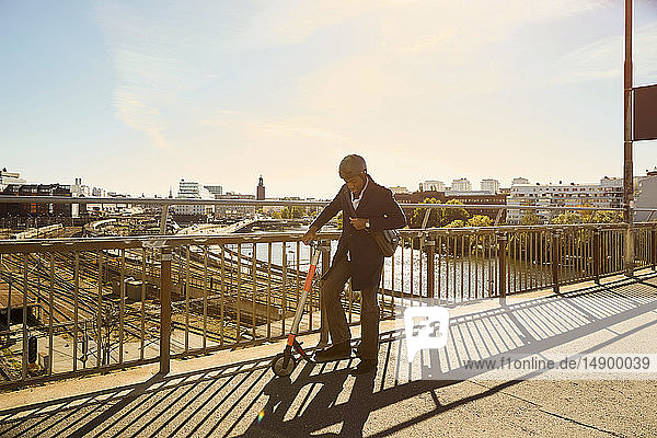 In voller Länge stehender junger Mann mit elektrischem Schieberoller an einem Geländer auf einer Brücke in der Stadt