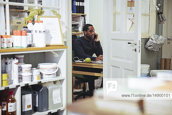 Selbstbewusster junger männlicher Besitzer,  der am Schreibtisch im Möbelgeschäft sitzt und mit einem Smartphone telefoniert