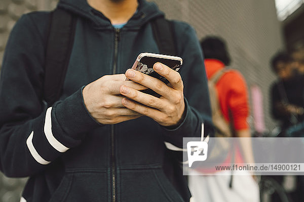 Mittelsektion eines jungen Mannes  der mit Freunden im Hintergrund auf einem Fußweg ein Mobiltelefon benutzt