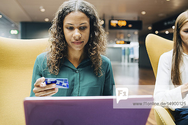 Zuversichtliche Geschäftsfrau mit Kreditkarte  während sie den Laptop ihres Kollegen in der Flughafen-Lobby benutzt