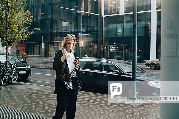 Geschäftsfrau benutzt Smartphone beim Gehen auf dem Bürgersteig gegen Gebäude in der Stadt