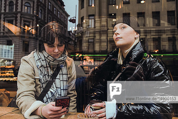 Nachdenkliche junge Frau  die durch ein Glasfenster schaut  während ein Freund im Winter im Café ein Mobiltelefon benutzt