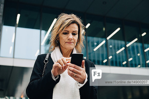 Niedrigwinkelansicht einer Geschäftsfrau mit blonden Haaren  die ein Smartphone benutzt  während sie vor einem modernen Gebäude in der Stadt steht