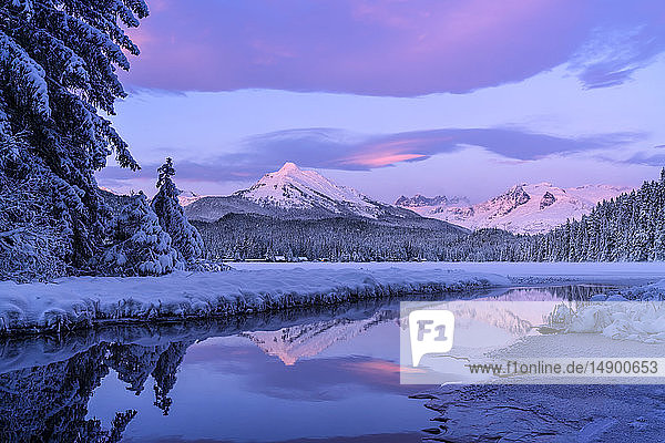 Alpenglow colours the snow-covered coastal range mountains Mendenhall Glacier and Mendenhall towers are reflected in the open water along the edge of Auke Lake as the sun gets ready to set; Alaska United States of America