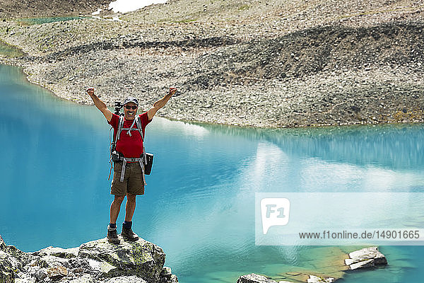 Male hiker standing on a large rocky area with a colourful alpine lake and rocky shoreline in the background with arms up in the air; British Columbia Canada